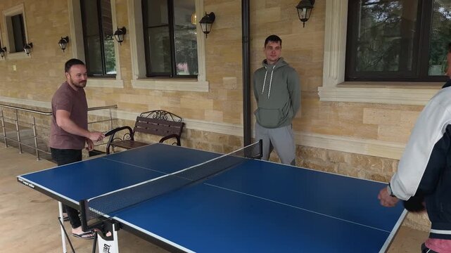 Observer catches stray ping pong ball and hands it back as beginner serves during friendly table tennis game on covered house terrace with stone wall, windows, porch lamps and relaxed family vibe