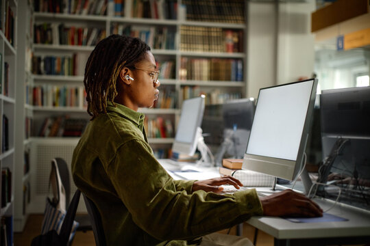 African American student studying on desktop computer in library, wearing earbuds and researching coursework
