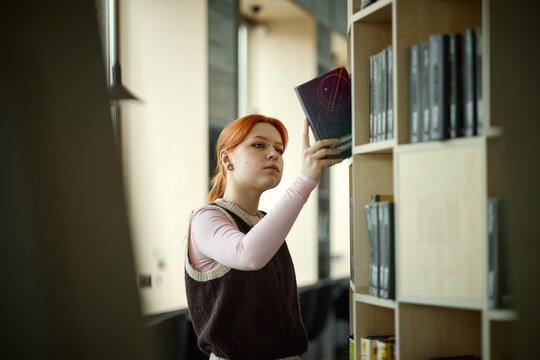 Caucasian woman selecting book from library shelf, studying and researching for college assignment

