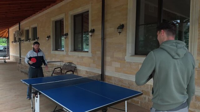 Smiling father and adult son share playful table tennis exchange on covered veranda, joking during casual home recreation as family friend observes near railing, blue table and warm wooden roof