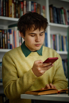 Vertical shot of Caucasian male student using smartphone while studying with open book in library
