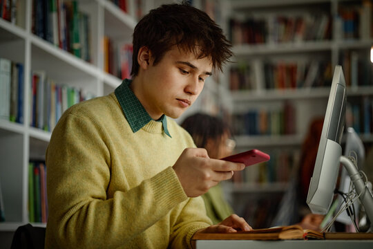 Student using smartphone while studying at desktop computer in library, multitasking with digital learning tools
