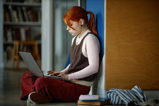 Caucasian student studying on laptop while sitting on floor in library, focusing on online learning and academic work
