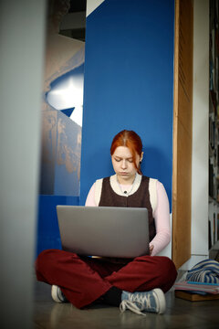 Vertical shot of Caucasian student studying on laptop while sitting on floor in library, focusing on online learning and academic research
