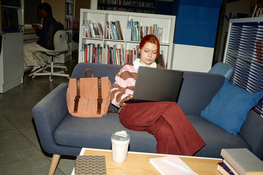 Young adult Caucasian woman studying on laptop in library lounge, researching coursework and online learning. Useful for education marketing, student life, academic services
