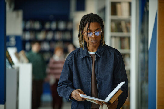 Medium shot of Black student reading book while walking through library, studying for education goals
