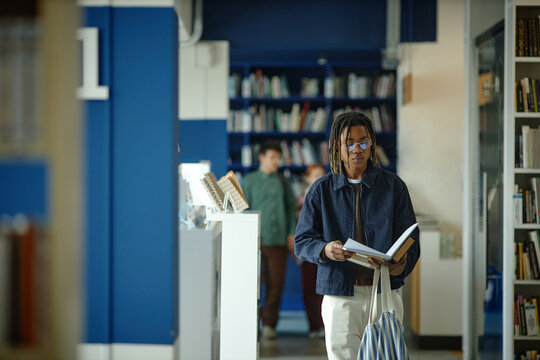 Young Black male student reading book while walking through library aisle, studying independently for academic success
