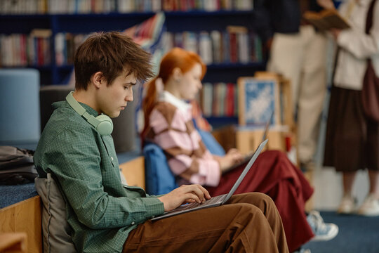 Caucasian student studying on laptop in library, wearing headphones and focusing on task while female classmate working in background. Useful for education, e learning, student life
