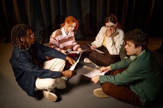 Young adult drama students rehearsing script together while sitting on floor, discussing lines and preparing performance
