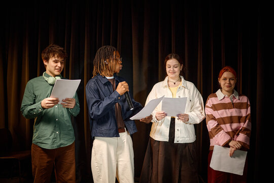Multiethnic students presenting scripts on stage, speaking into microphone during college performance rehearsal. Useful for education marketing, youth programs, teamwork campaigns
