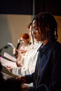 Vertical shot of Black student reading script during college presentation while classmates speaking into microphone, useful for education, public speaking, youth learning
