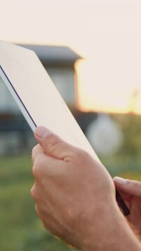 Businessman promptly carries out online deal to sell crop. Close up of farmers hands using digital computer and searches for information in tablet in agricultural field with plants in windy weather on
