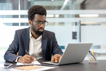 Obraz premium Young man wearing business suit and glasses, working diligently at a modern office desk, typing on his laptop and simultaneously writing down important notes in a professional workspace