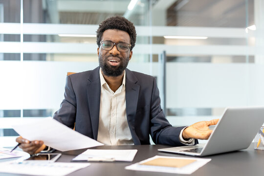 Professional man showing confusion and frustration, holding documents and gesturing towards a laptop while experiencing work stress or a challenging problem in a modern office setting
