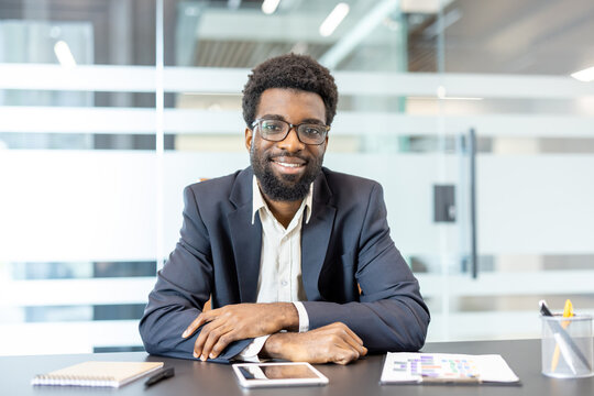 Confident young professional man sitting at desk in a bright contemporary office setting, smiling and looking directly at camera, representing concepts of success, career, and corporate leadership