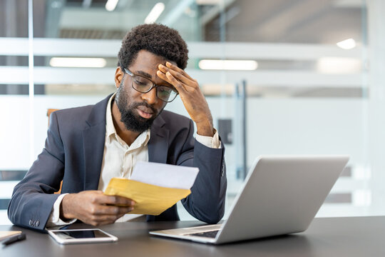 Stressed businessman in a modern office receiving bad news in a letter, experiencing worry, debt, and loss while looking at a laptop and smartphone on his desk