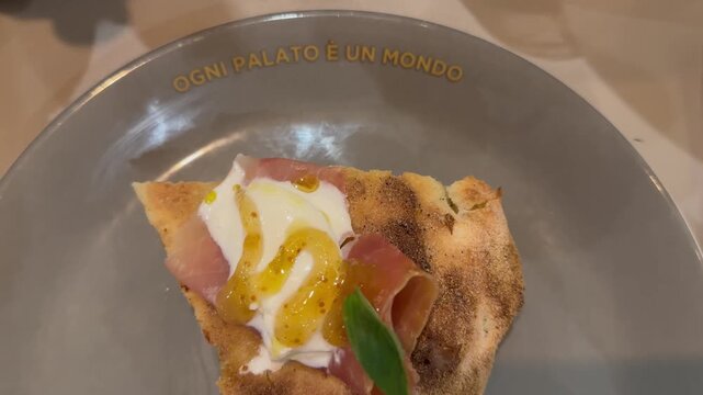 a delicious slice of pizza with stracciata, ham, and fig jam on a plate that says: 'every palate is a world'