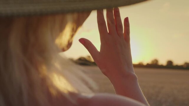 Woman wearing straw hat looks at golden sunset through her fingers in wheat field.