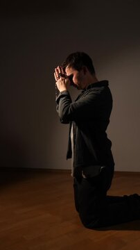 Vertical video man kneeling in contemplation, hands clasped around rosary beads, expressing faith and devotion while meditating in a moment of spiritual solitude and hope on a dark background