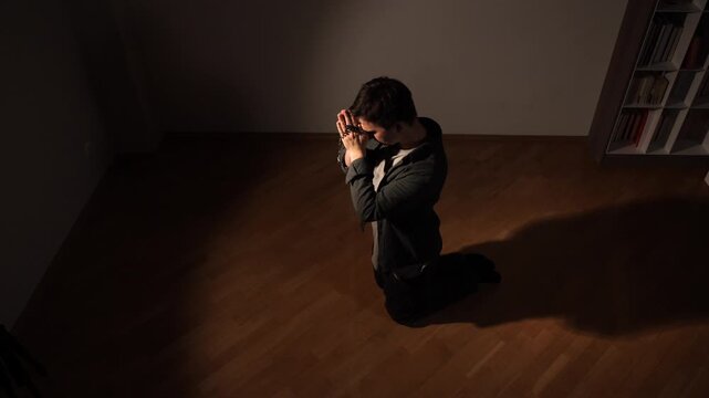 Man kneeling in dim room holding rosary beads, praying in solitude, quiet moment of faith, reflection and hope during spiritual meditation and seeking solace