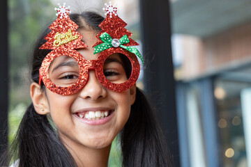 Indian female child smiling and wearing red glitter Christmas-tree glasses at cafe windows © wavebreak3