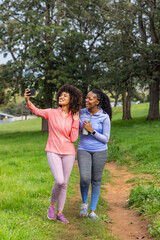 Fototapeta premium Diverse female friends walking in sportswear on park path taking selfie with phone holding bottle
