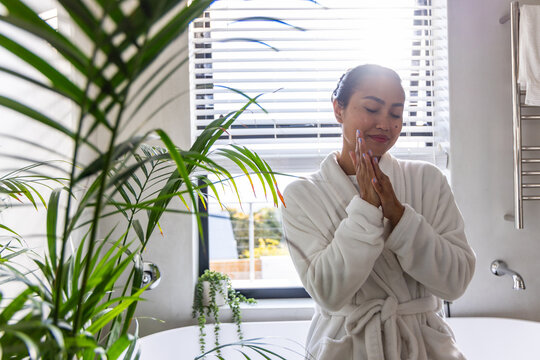 Asian woman doing skincare in bathroom wearing robe by freestanding tub with plants, copy space