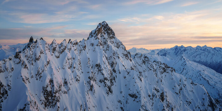Aerial view of the snow-covered jagged peaks of Chugach State Park under a soft sunset sky with golden light hitting the mountain ridges in Anchorage, Alaska, United States.