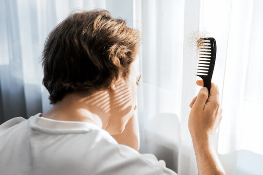 Over shoulder view of a stressed man looking down at a comb full of lost hair, representing male hair loss, testosterone issues, and alopecia symptoms during grooming.