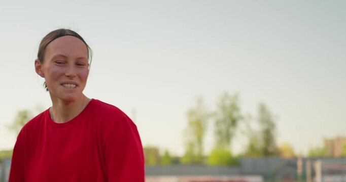 Young female runner in a red shirt training on an outdoor stadium track at sunset, smiling in profile with bleachers and field behind her