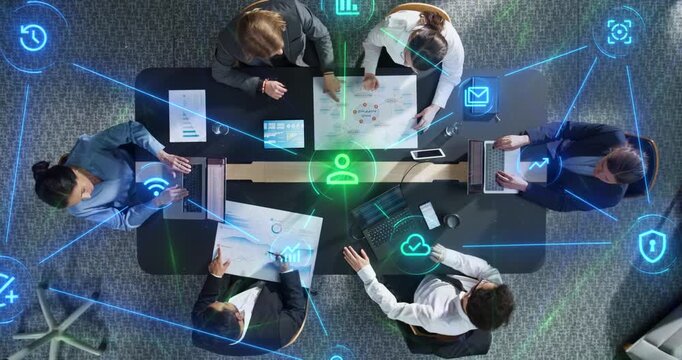 Top Down View of Diverse Office Team Strategizing Around Meeting Table With Laptops and Reports as Linked Visualization Layer Appears, Glowing Tech Icons Connectivity, Productivity, Cybersecurity.