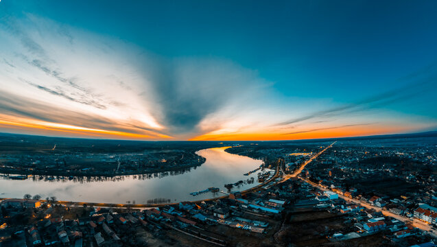Aerial view of the Sava river winding through the urban landscape and illuminated streets at sunset under a dramatic sky in Sremska Mitrovica, Vojvodina, Serbia.