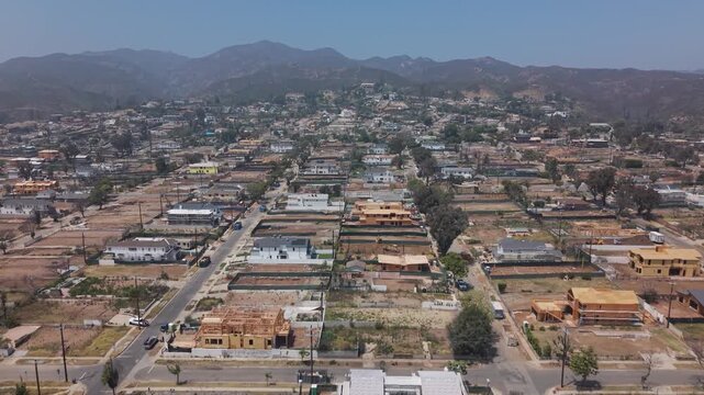 Hillside neighborhood with wildfire damage, empty lots, scattered surviving homes, dry mountain range, sparse trees, and ongoing reconstruction under a sunny blue sky. California, USA.Aerial wide shot