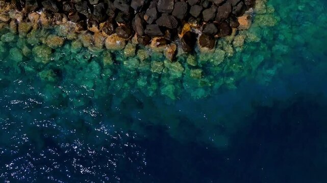Aerial view shows volcanic rock breakwater and clear blue ocean meeting on the coast