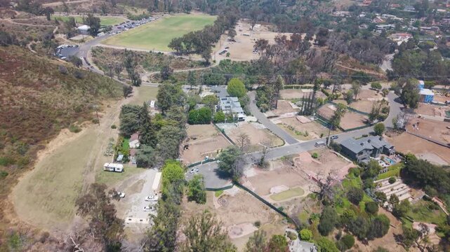 Suburban area with wildfire aftermath showing burned trees, empty lots, scattered surviving structures, a green sports field, and dry hillside vegetation. California, USA. Aerial tilt up wide shot.