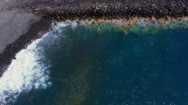 Waves wash against rocky breakwater and clear blue sea along volcanic stone shore