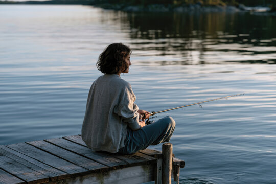 A woman sits peacefully on a dock, engaged in fishing while appreciating the calm water surroundings, embodying a moment of solitude and connection to nature.