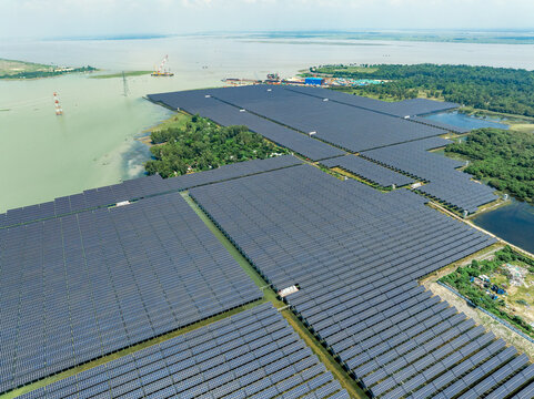 Aerial view of a massive solar power plant with rows of photovoltaic panels situated along the riverbank near industrial activity in Sirajganj, Rajshahi Division, Bangladesh.