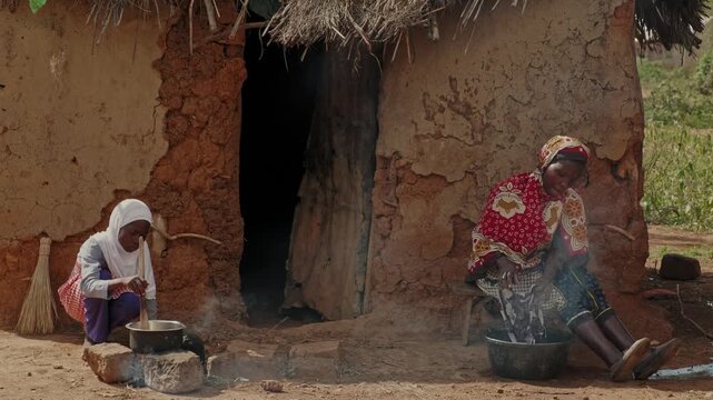 African mother washing clothes beside traditional mud houses while a child helps prepare food nearby. Authentic rural village life scene under bright blue sky in countryside Africa.