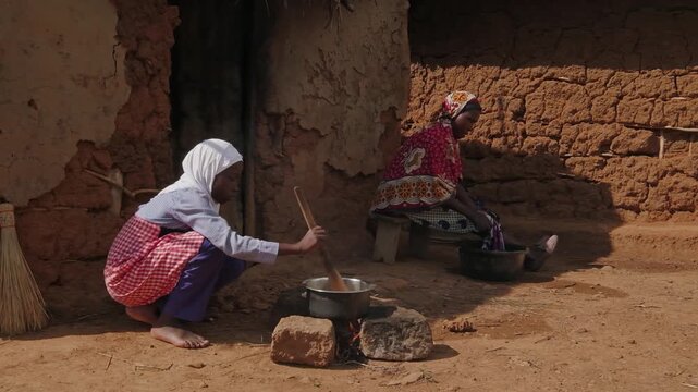 African mother washing clothes beside traditional mud houses while a child helps prepare food nearby. Authentic rural village life scene under bright blue sky in countryside Africa.