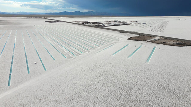 Aerial view of Salinas Grandes salt flats featuring turquoise lithium evaporation ponds under a dramatic dark sky and distant mountains in Tumbaya, Jujuy, Argentina.