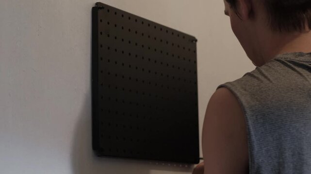 Medium rear shot of a young man using a screwdriver to install a black pegboard on a white wall in an indoor workspace