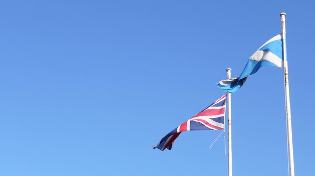 Low angle view of the United Kingdom Union Jack flag and the Scottish Saltire flag waving on flagpoles against a clear blue sky background