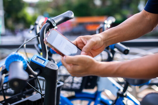 A close-up shot of hands holding a smartphone to scan a QR code on a blue shared bicycle's handlebars, unlocking it for a ride on a blurred city street.