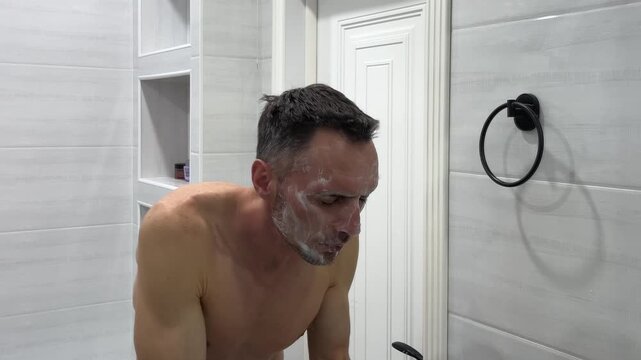 A shirtless athletic man washes himself over a sink with soapy foam and rinses it off with water, performing his daily morning hygiene procedures