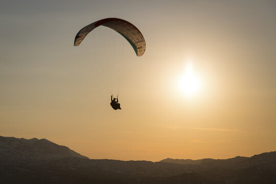 Aerial view of a paraglider in silhouette soaring over the snow-covered peaks of Hoher Dachstein during a golden sunset in Liezen District, Styria, Austria.