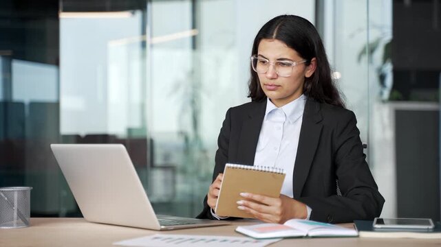 Young businesswoman listens to online webinar on laptop and writes notes in notebook at workplace in modern office. Focused professional learning remotely, engaged in business training.