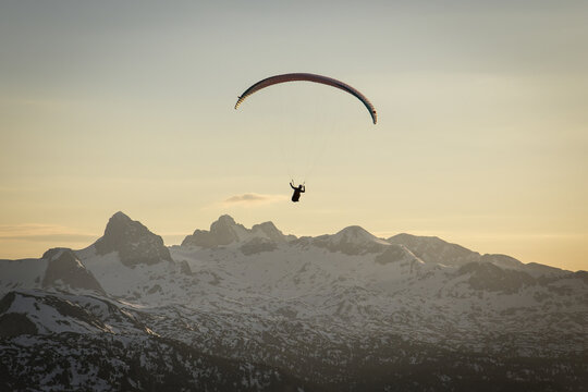 Aerial view of a paraglider soaring over the snow-capped peaks of Hoher Dachstein during a golden sunset in Liezen District, Styria, Austria.