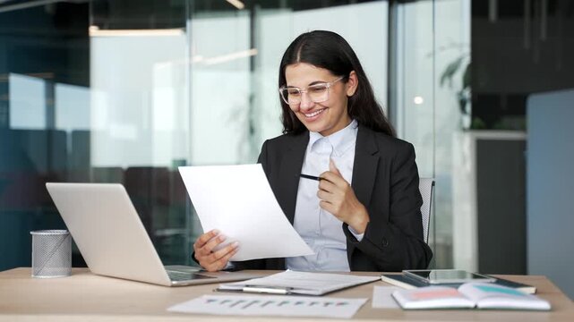 Smiling businesswoman reviewing financial documents and satisfied with results while sitting in business office. Happy young female financier rejoices at positive indicators, celebrates success.