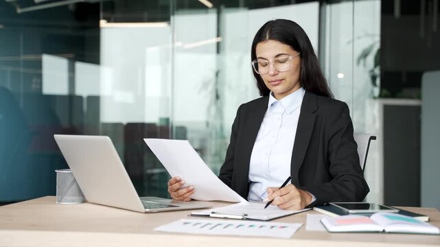 Female accountant filling out documents or tax form with a pen while sitting at desk at workplace in business office. Businesswoman reviews paperwork, calculates expenses and manages company finances.